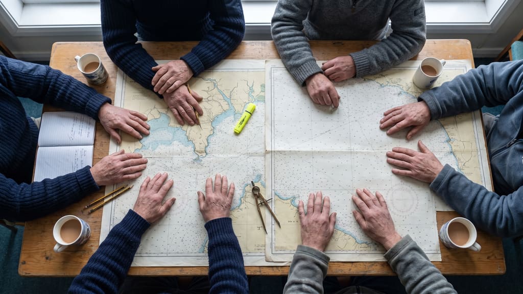 Six people around a table with a nautical chart spread out between them, seen from above.