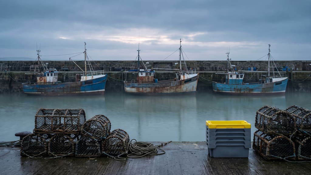 Three small trawlers moored against the harbour wall at dusk, with creels and rope stacked on the quay.
