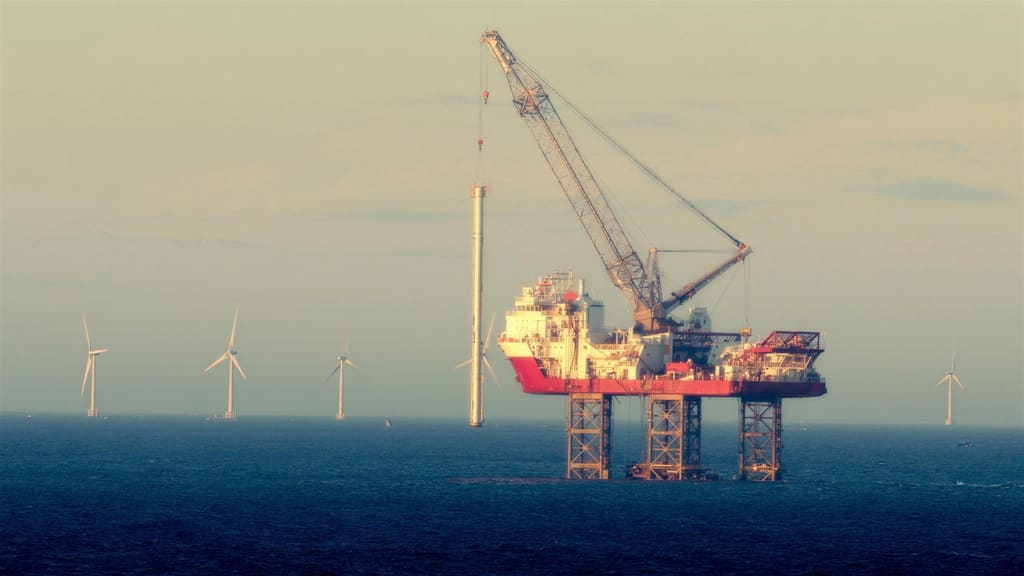 A jack up installation vessel holding a monopile upright on its crane at sea, with turbines already standing behind it.