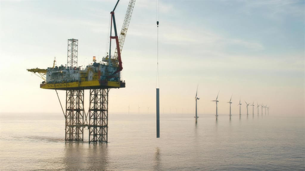 A jack up vessel raised clear of the water lowering a monopile over the side, with a row of turbines running off into the haze.